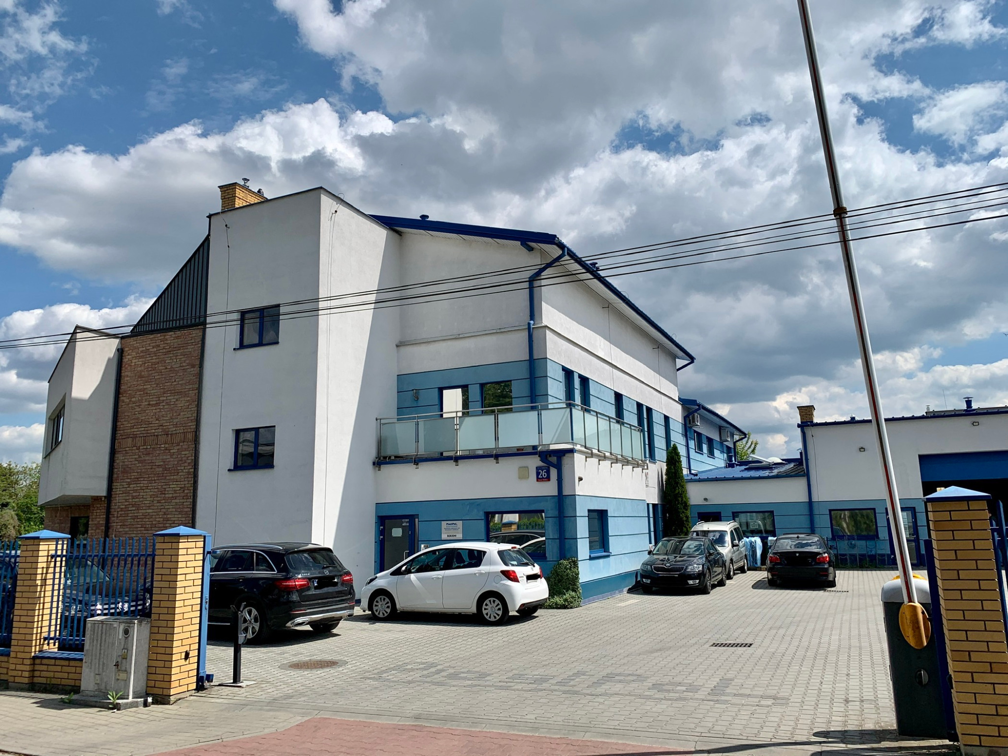 Dwupiętrowy budynek biurowy z białymi ścianami i niebieskimi akcentami, zaparkowane samochody na brukowanym parkingu, brama wjazdowa po prawej stronie.