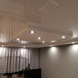 Build in Trust - Błyszczący, kremowy sufit napinany z kwadratową zabudową oświetleniową z sześcioma punktami LED, widok na szare ściany i firany z koronkowym wykończeniem.