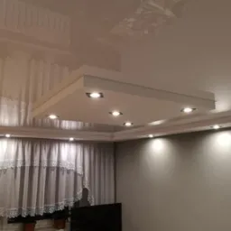 Błyszczący, kremowy sufit napinany z kwadratową zabudową oświetleniową z sześcioma punktami LED, widok na szare ściany i firany z koronkowym wykończeniem.