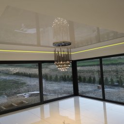 Build in Trust - Błyszczący, biały sufit napinany z podświetleniem LED i nowoczesnym żyrandolem w przestronnym salonie z dużymi oknami przesuwnymi na ogród.