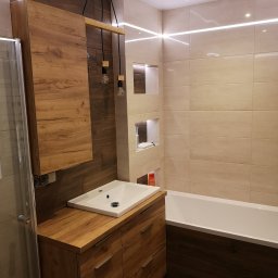 Build in Trust - Nowoczesna łazienka z drewnianymi akcentami: szafka pod umywalką z szufladami, wisząca szafka, drewniana lampa z żarówkami Edisona, beżowe płytki, wanna obudowana drewnem i oświetlenie LED na suficie.