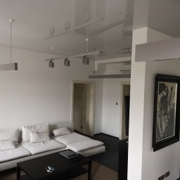 Build in Trust - Nowoczesny salon z białymi ścianami, sofą modułową i błyszczącym sufitem napinanym, oświetlonym przez podwieszane lampy szynowe i wiszące lampy listwowe.