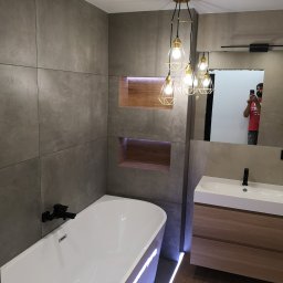 Build in Trust - Nowoczesna łazienka z szarymi płytkami wielkoformatowymi, podświetlaną wanną, drewnianymi wnękami w ścianie, geometryczną lampą wiszącą i odbiciem osoby robiącej zdjęcie w lustrze nad umywalką.