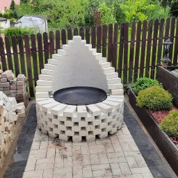 Build in Trust - Ogród z murowanym grillem z białej cegły, z czarnym rusztem w środku, otoczony kostką brukową i rabatami z roślinami, widok z góry.