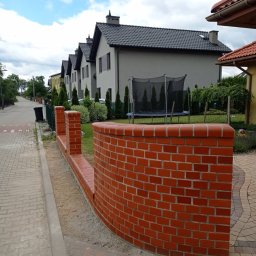 Build in Trust - Nowo wymurowany, zakrzywiony murek z czerwonej cegły, oddzielający posesję od chodnika, z widocznymi śladami zaprawy i piasku, z rzędem domów w tle.