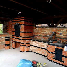 Build in Trust - Murowany grill ogrodowy z cegły klinkierowej z czarnymi akcentami, widoczne narzędzia i materiały budowlane, częściowo zadaszony.