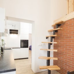 Build in Trust - Nowoczesna kuchnia z białymi szafkami, blatem w kolorze grafitu i spiralnymi schodami z drewnianymi stopniami prowadzącymi na antresolę z drewnianą balustradą, ściana z czerwonej cegły.