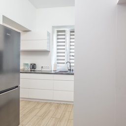 Build in Trust - Nowoczesna, jasna kuchnia z białymi szafkami, szarym blatem, srebrną lodówką Amica i drewnopodobną podłogą, widziana z perspektywy wejścia.