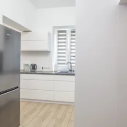 Nowoczesna, jasna kuchnia z białymi szafkami, szarym blatem, srebrną lodówką Amica i drewnopodobną podłogą, widziana z perspektywy wejścia.