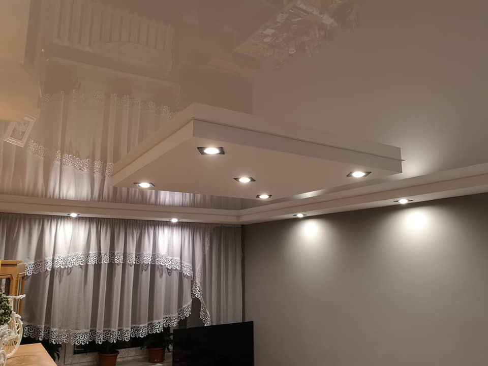 Błyszczący, kremowy sufit napinany z kwadratową zabudową oświetleniową z sześcioma punktami LED, widok na szare ściany i firany z koronkowym wykończeniem.