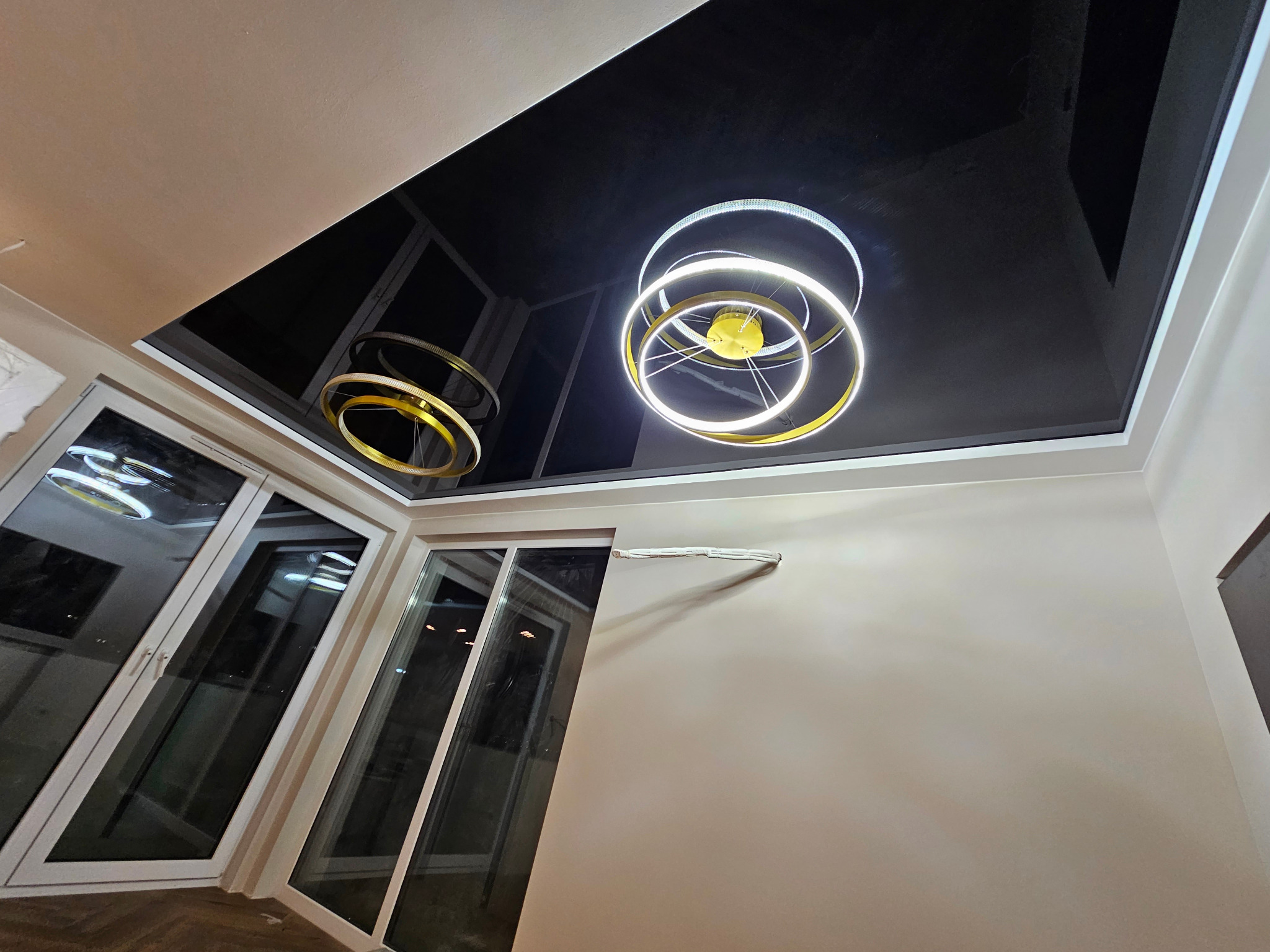 Czarny, lustrzany sufit napinany z nowoczesnym, okrągłym oświetleniem LED w nowocześnie wykończonym salonie z dużymi oknami balkonowymi.