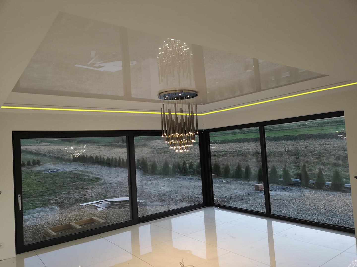 Błyszczący, biały sufit napinany z podświetleniem LED i nowoczesnym żyrandolem w przestronnym salonie z dużymi oknami przesuwnymi na ogród.