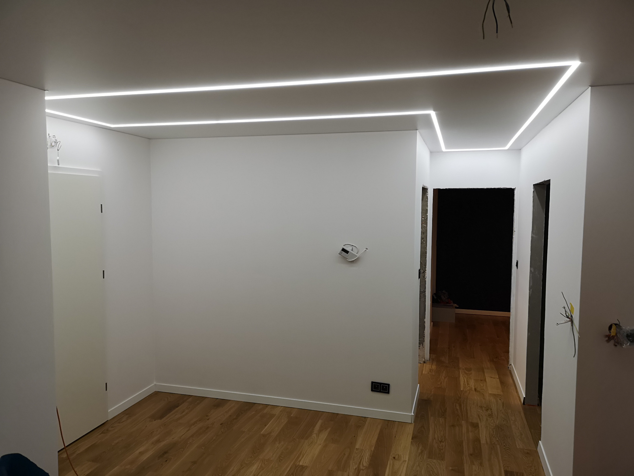 Wnętrze po remoncie, z drewnianą podłogą i białymi ścianami, oświetlone listwami LED w suficie, widoczne niedokończone prace elektryczne.
