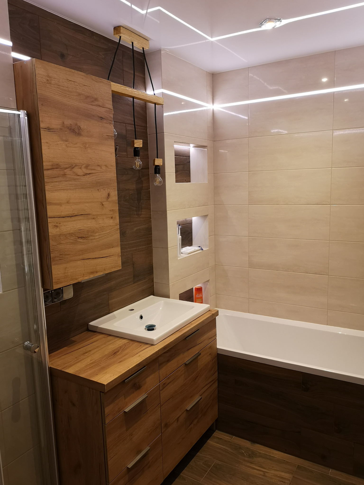 Nowoczesna łazienka z drewnianymi akcentami: szafka pod umywalką z szufladami, wisząca szafka, drewniana lampa z żarówkami Edisona, beżowe płytki, wanna obudowana drewnem i oświetlenie LED na suficie.