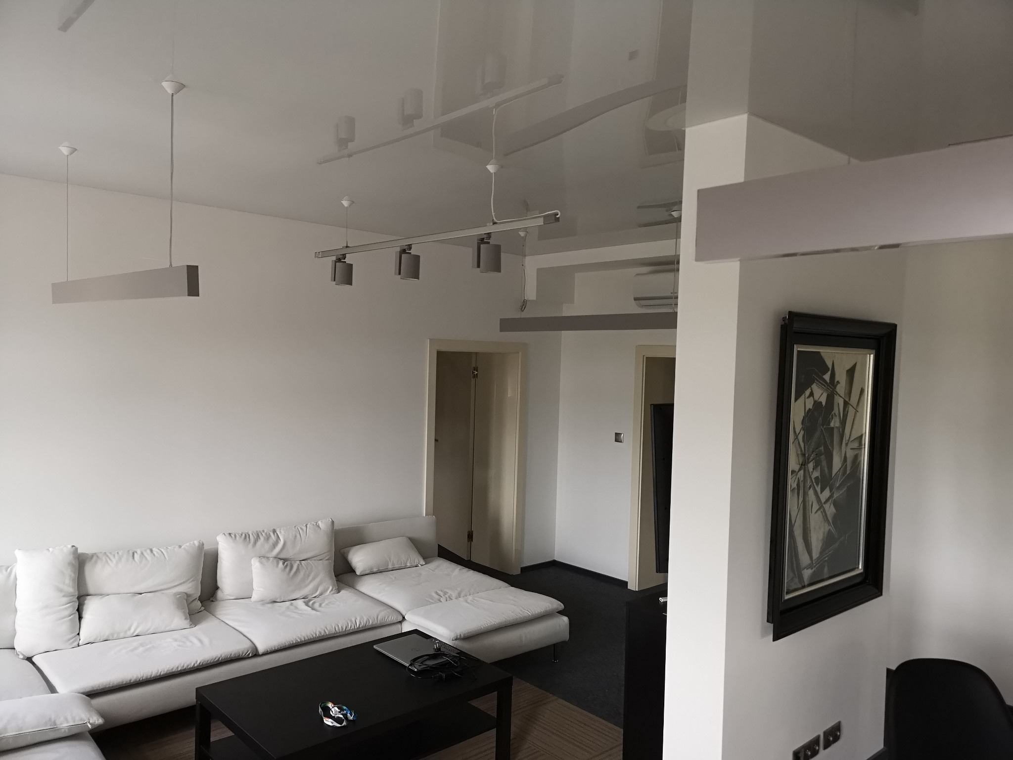 Nowoczesny salon z białymi ścianami, sofą modułową i błyszczącym sufitem napinanym, oświetlonym przez podwieszane lampy szynowe i wiszące lampy listwowe.