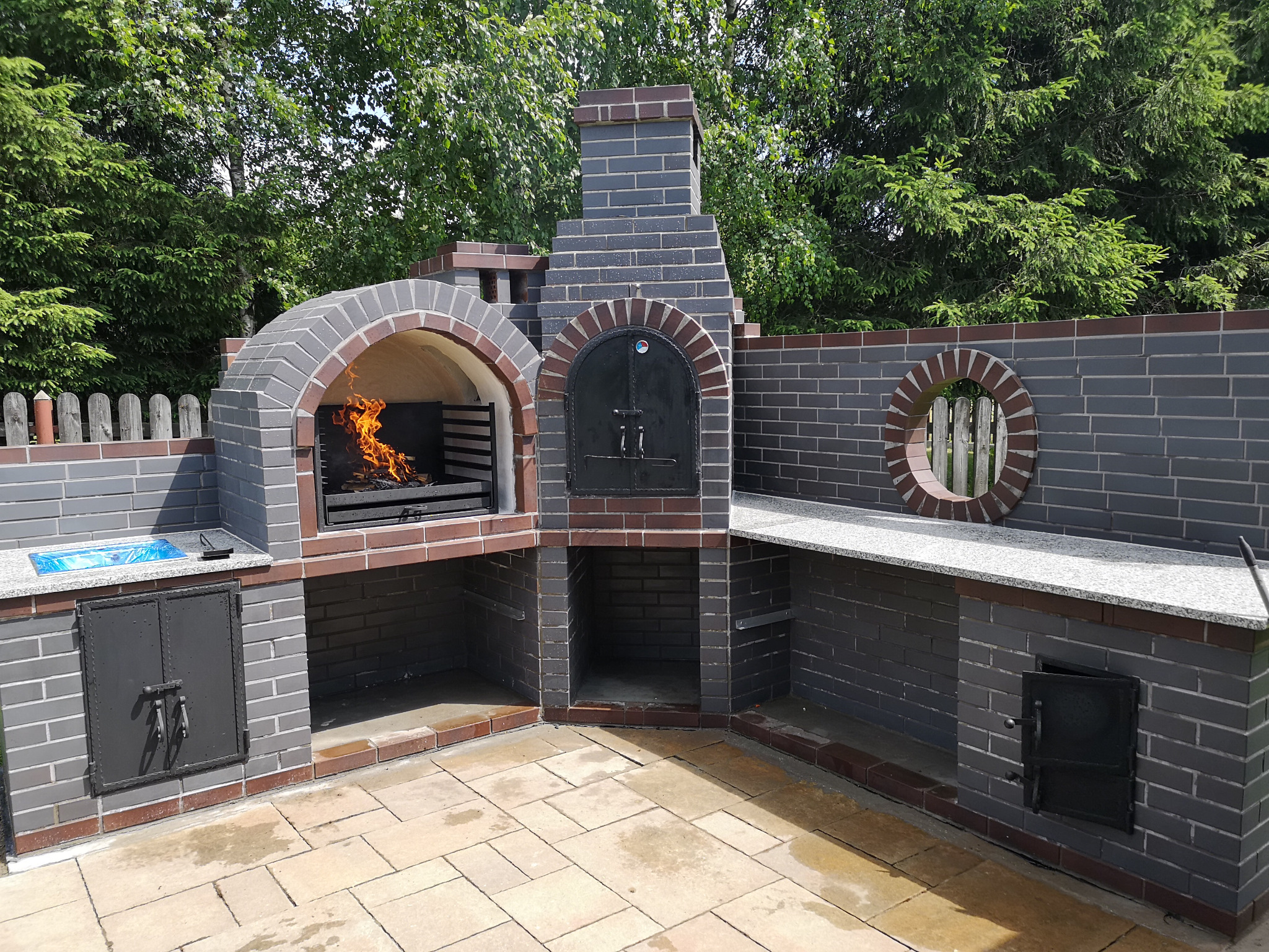 Murowany grill ogrodowy z paleniskiem wypełnionym płomieniami, w tle zielone drzewa.