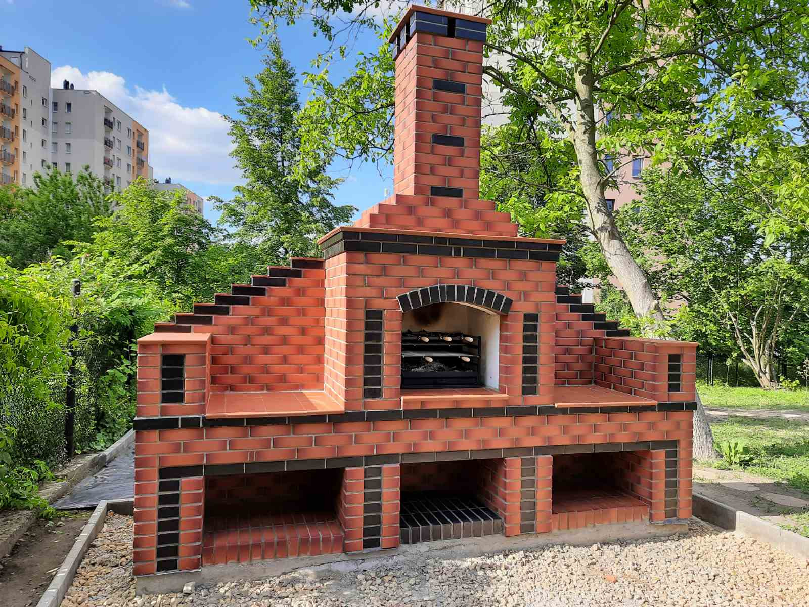 Murowany grill ogrodowy z czerwonej cegły z czarnymi akcentami, z rusztem i kominem, na tle zieleni i bloków mieszkalnych.