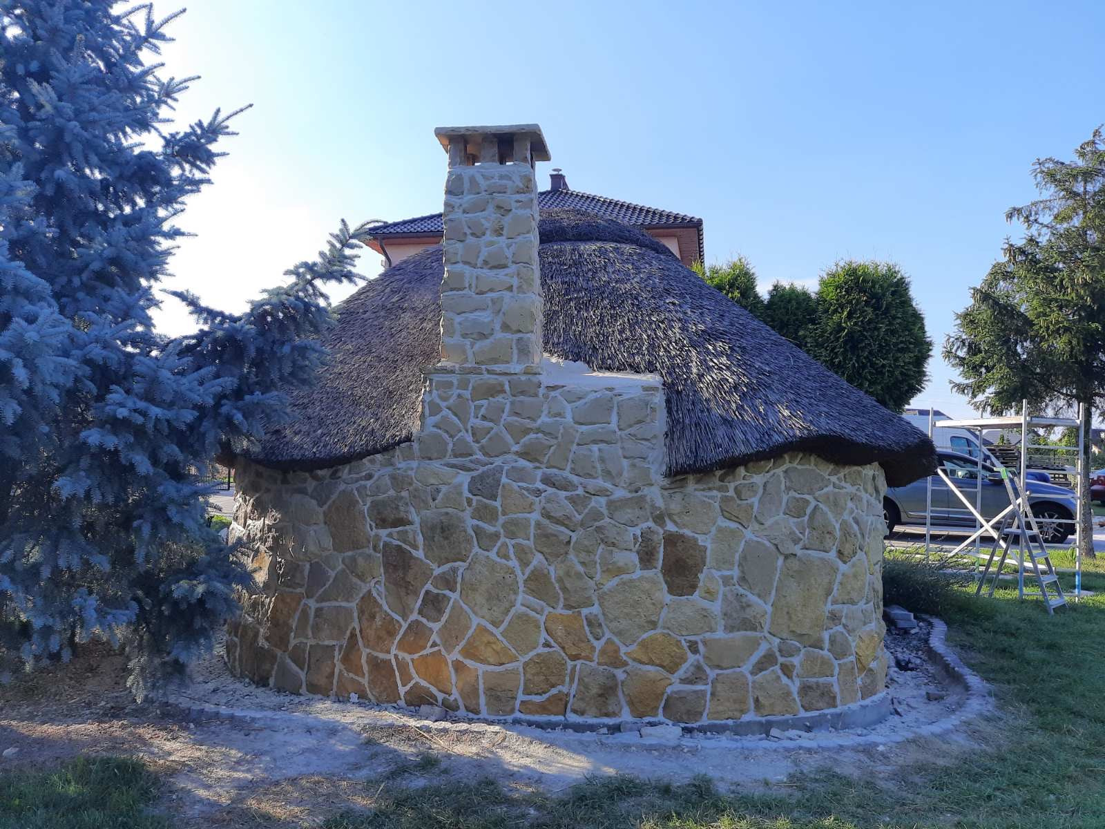 Okrągła altana z kamienną elewacją, kryta strzechą i z kamiennym kominem, obok rośnie srebrny świerk.