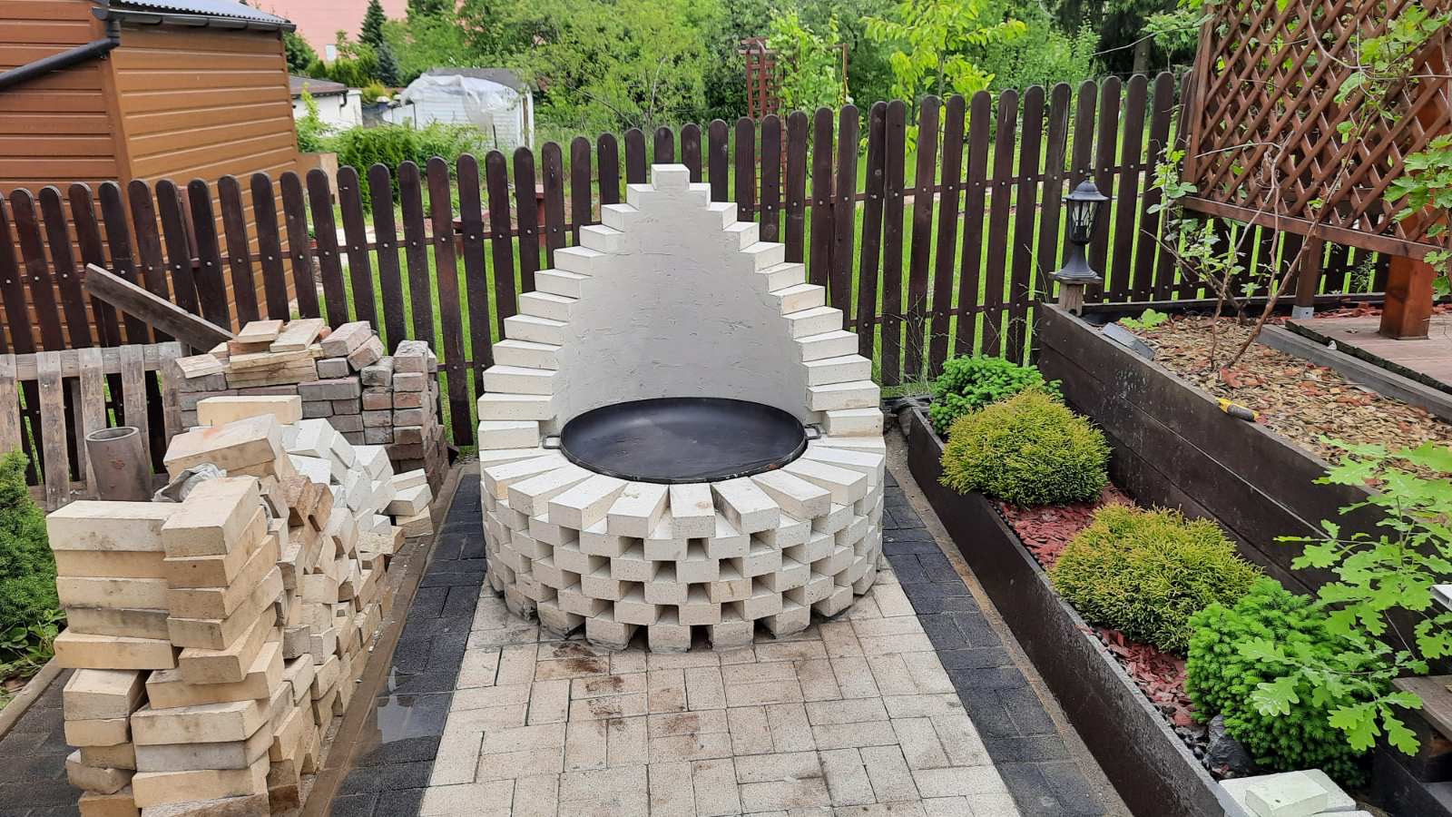 Ogród z murowanym grillem z białej cegły, z czarnym rusztem w środku, otoczony kostką brukową i rabatami z roślinami, widok z góry.