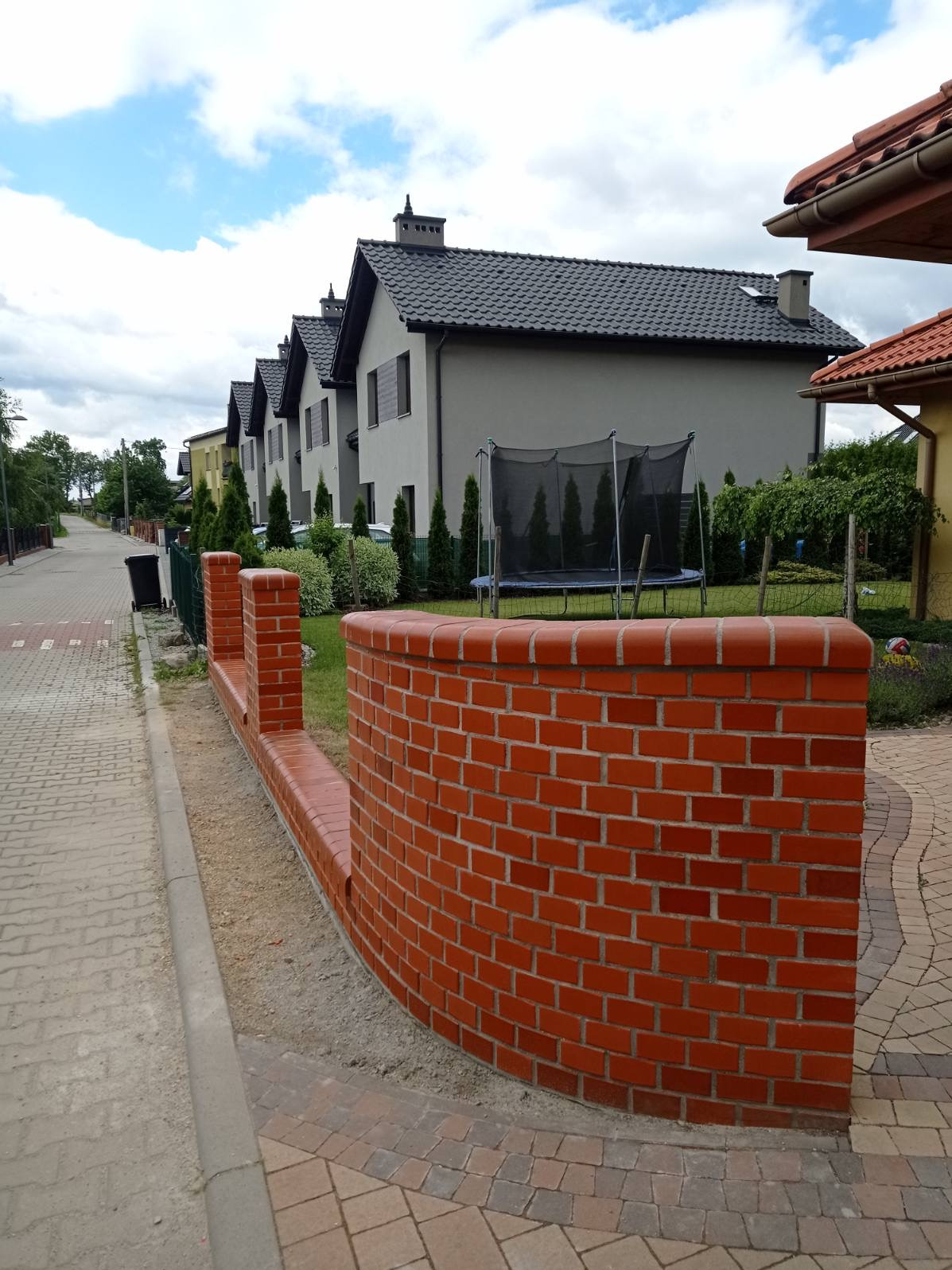 Nowo wymurowany, zakrzywiony murek z czerwonej cegły, oddzielający posesję od chodnika, z widocznymi śladami zaprawy i piasku, z rzędem domów w tle.