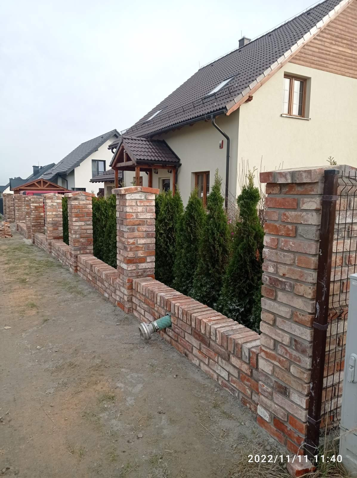 Nowo wybudowany ceglany mur z filarami, z widoczną zieloną rurą odpływową i tujami w tle, na tle domów jednorodzinnych.