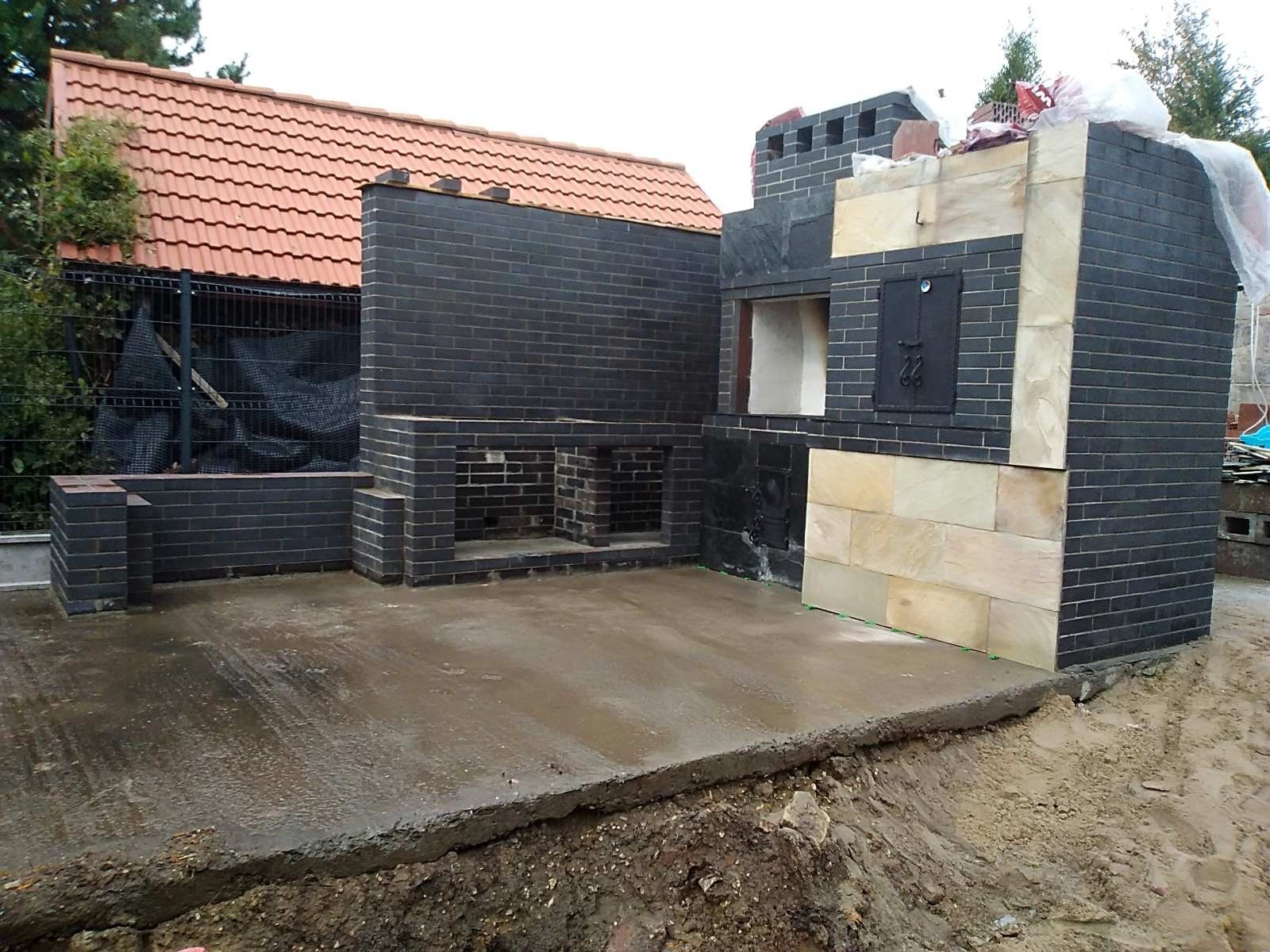 Nowo wybudowany, czarny, ceglany grill z wędzarnią i piecem chlebowym, z elementami piaskowca, na wilgotnym betonowym tarasie w Dąbrowie Górniczej.