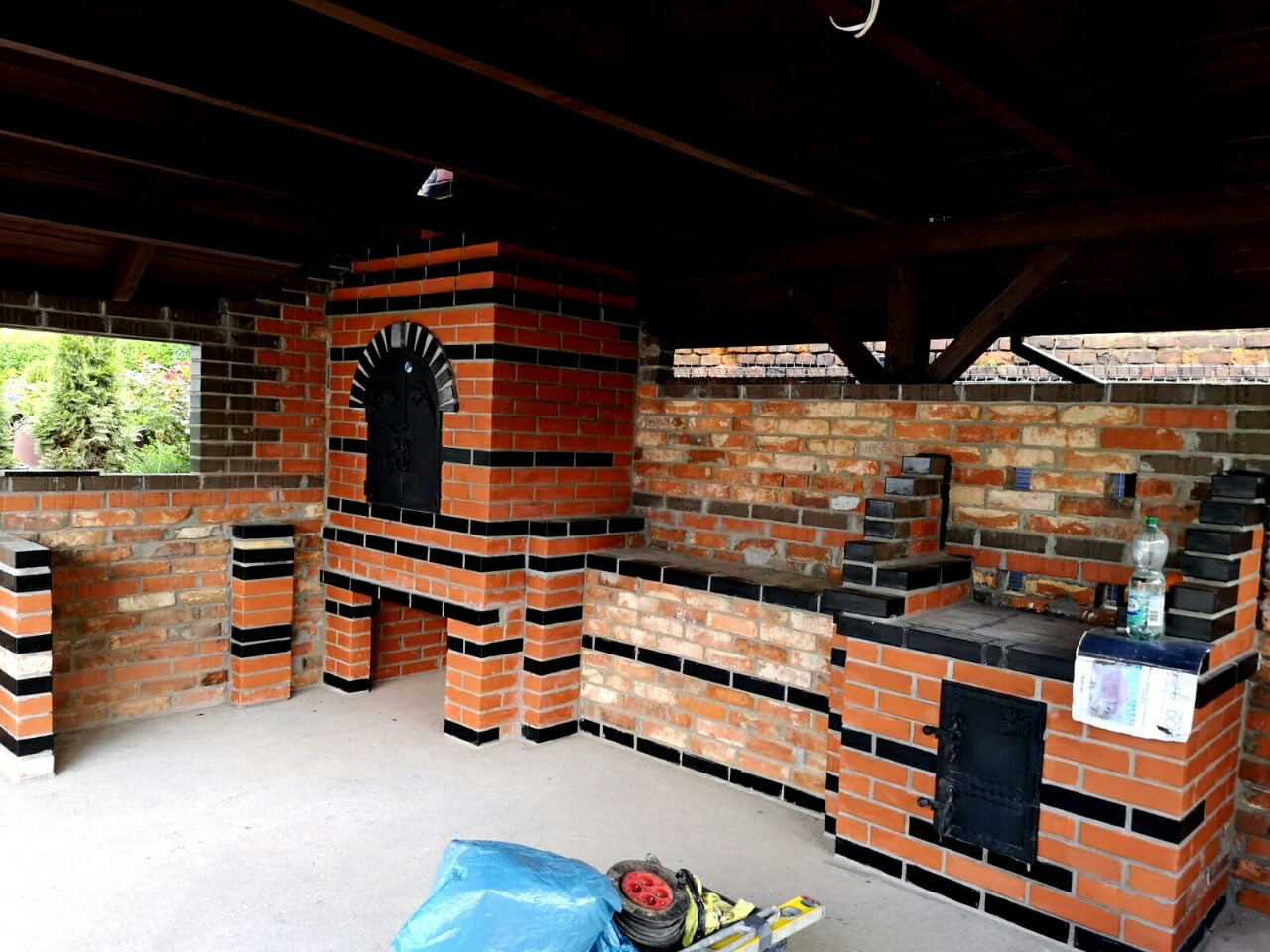 Murowany grill ogrodowy z cegły klinkierowej z czarnymi akcentami, widoczne narzędzia i materiały budowlane, częściowo zadaszony.