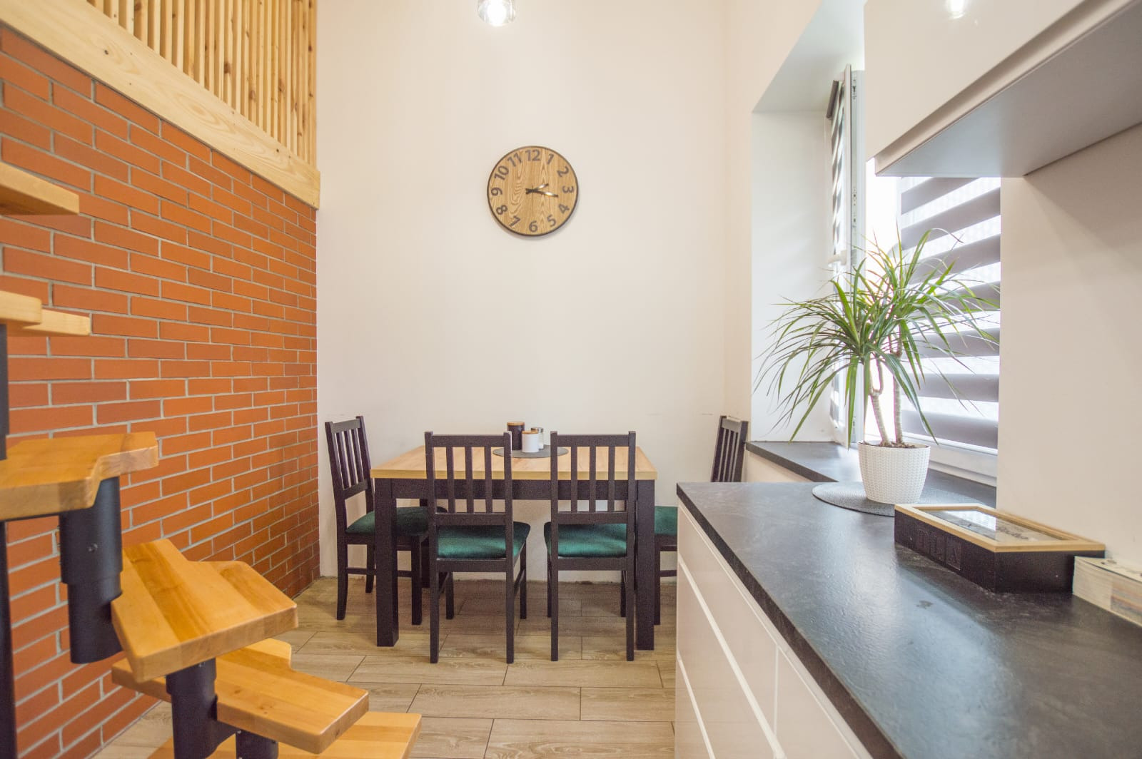 Jasne wnętrze z ceglaną ścianą, drewnianymi schodami, stołem z krzesłami z zielonymi poduszkami i zegarem na ścianie, uzupełnione rośliną doniczkową na blacie przy oknie.
