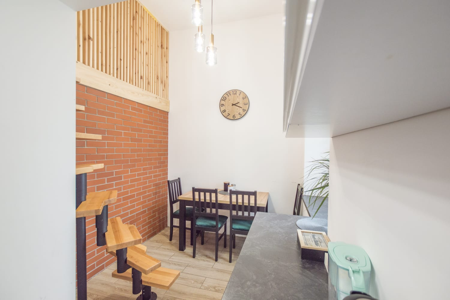 Wnętrze mieszkania po remoncie: ceglana ściana z drewnianą antresolą, drewniane schody spiralne, stół z krzesłami i zegar na ścianie, widok z perspektywy blatu kuchennego.