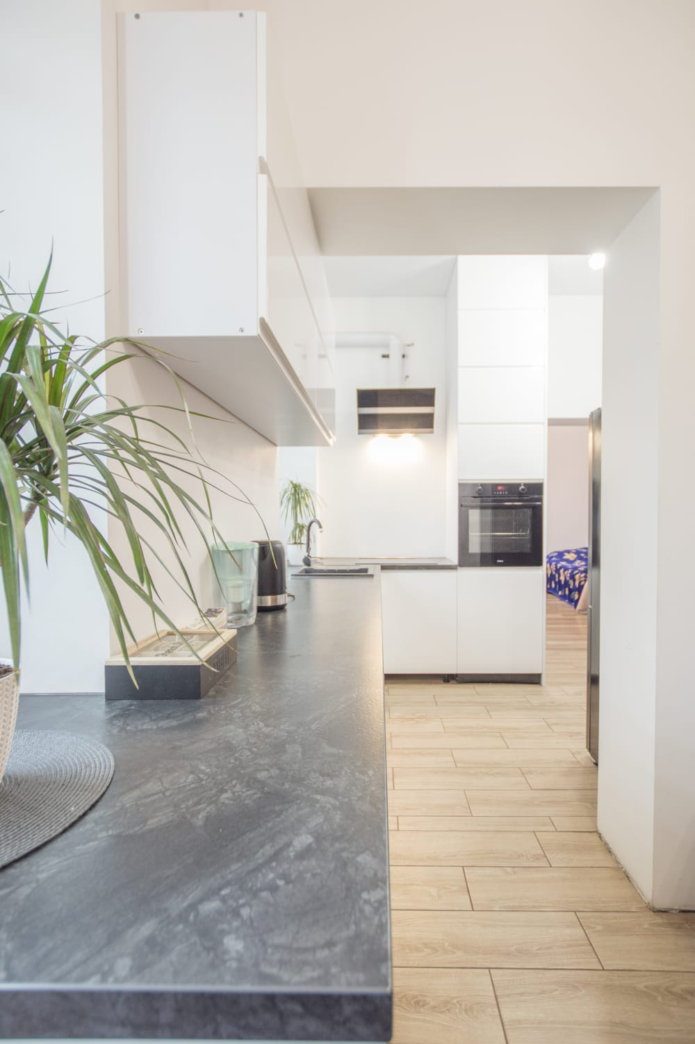 Nowoczesna kuchnia w Dąbrowie Górniczej z ciemnym blatem, białymi szafkami i drewnopodobną podłogą; widoczny czajnik elektryczny i roślina doniczkowa.