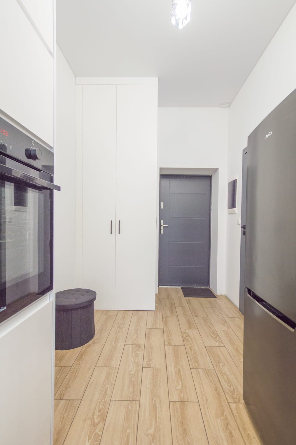 Wnętrze mieszkania: jasne płytki podłogowe imitujące drewno, białe szafy w zabudowie, szara pufa i drzwi wejściowe, widoczny fragment piekarnika i lodówki.