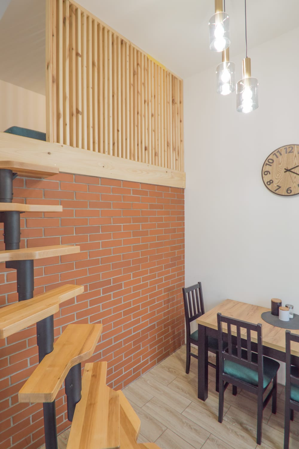 Wnętrze z ceglaną ścianą, drewnianymi schodami spiralnymi i antresolą z drewnianą balustradą, oświetlone wiszącymi lampami i z drewnianym zegarem na ścianie.