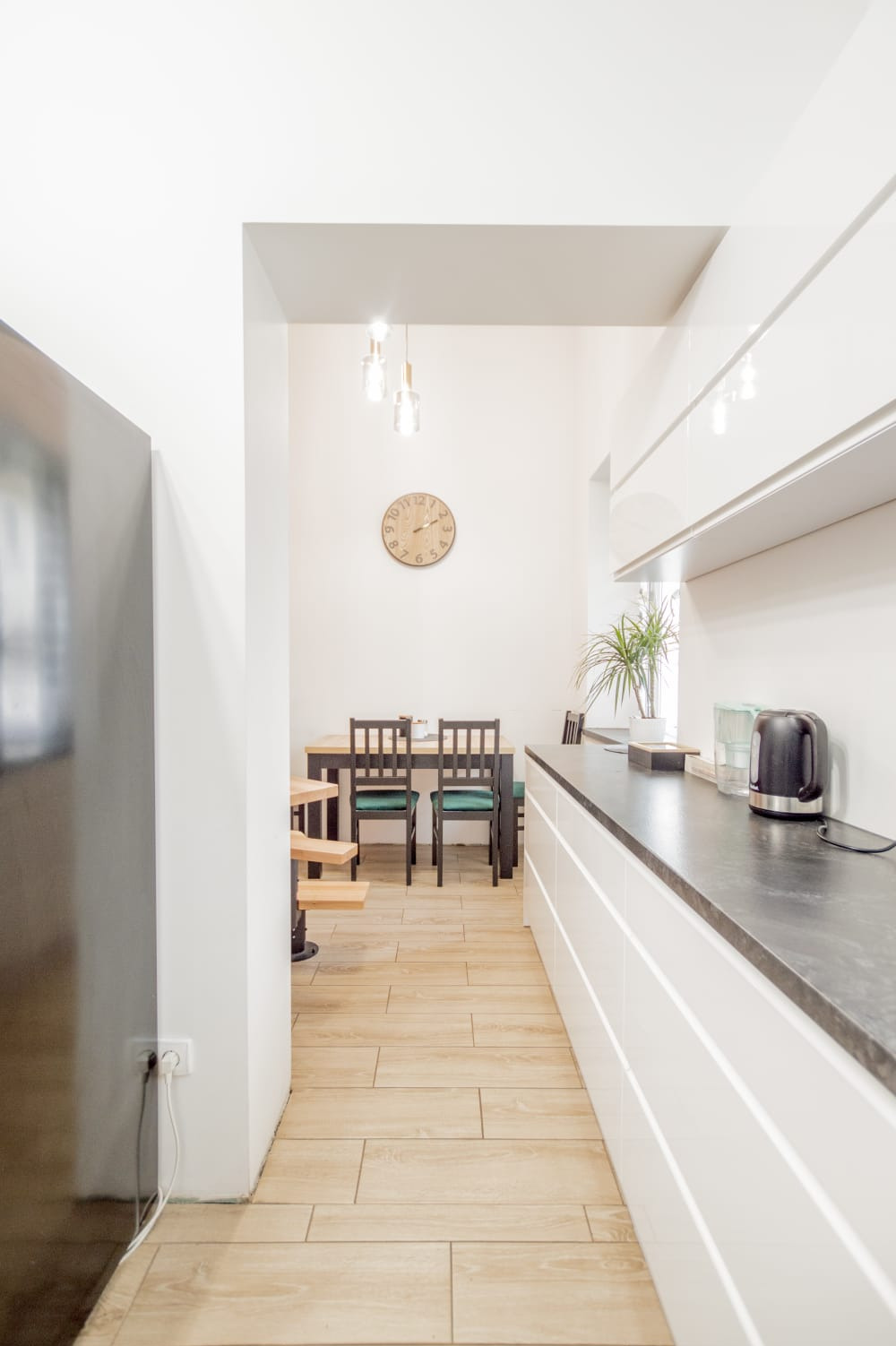 Jasna kuchnia z białymi szafkami, czarnym blatem, drewnianą podłogą imitującą deski i stołem z krzesłami w tle, widok z perspektywy wejścia.