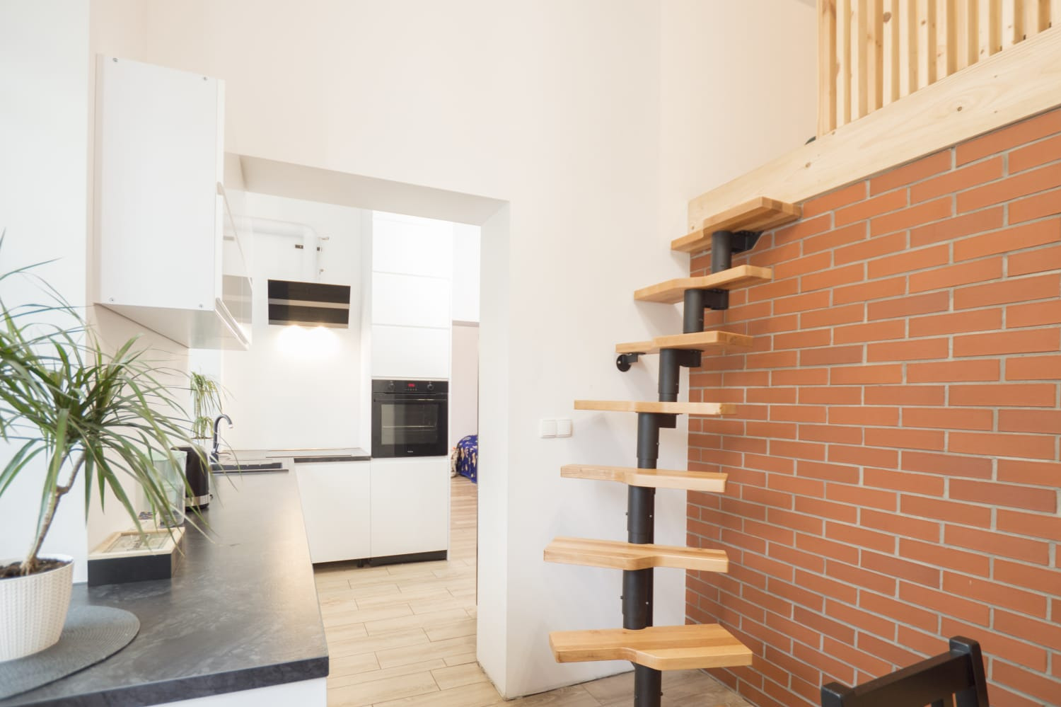 Nowoczesna kuchnia z białymi szafkami, blatem w kolorze grafitu i spiralnymi schodami z drewnianymi stopniami prowadzącymi na antresolę z drewnianą balustradą, ściana z czerwonej cegły.