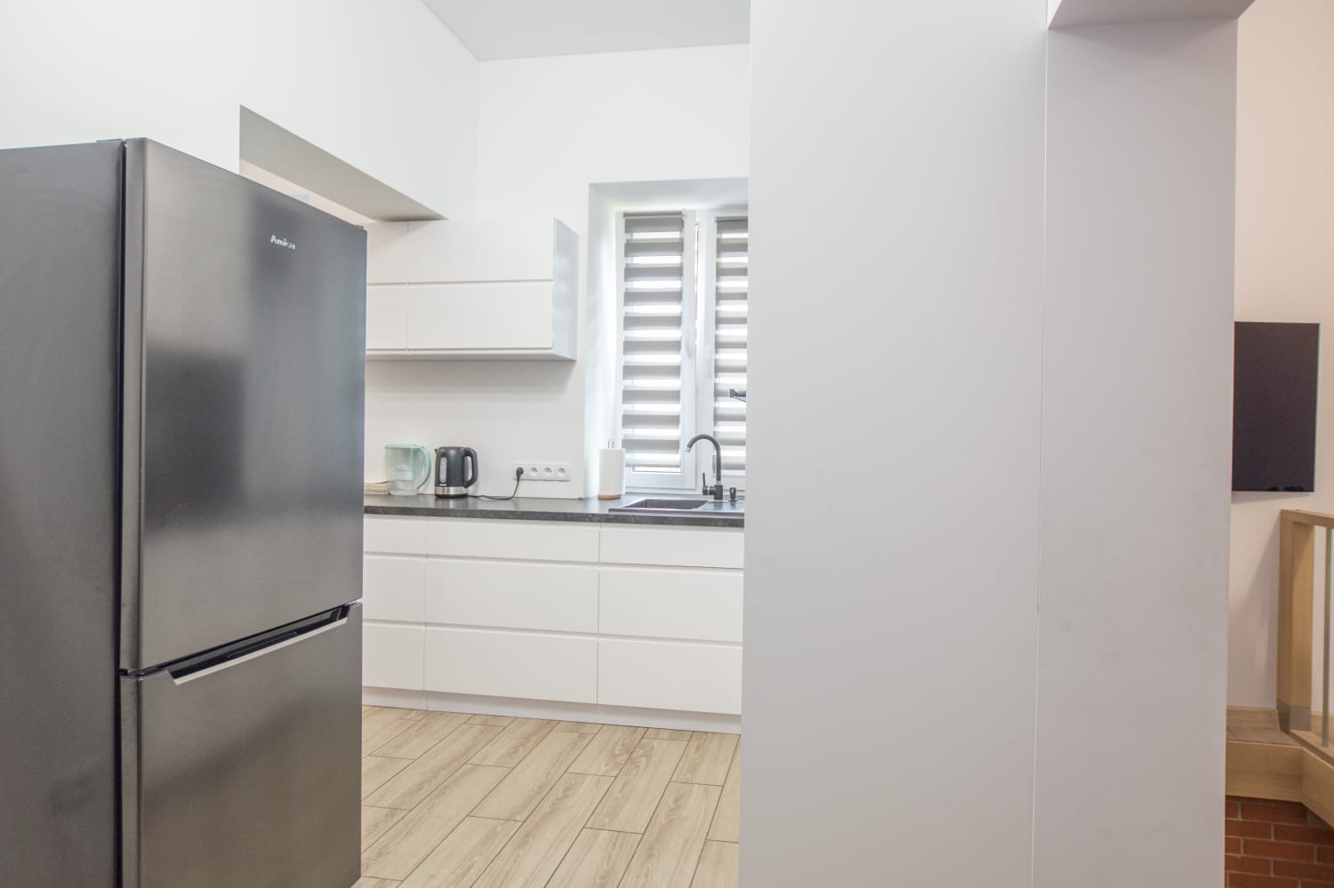 Nowoczesna, jasna kuchnia z białymi szafkami, szarym blatem, srebrną lodówką Amica i drewnopodobną podłogą, widziana z perspektywy wejścia.