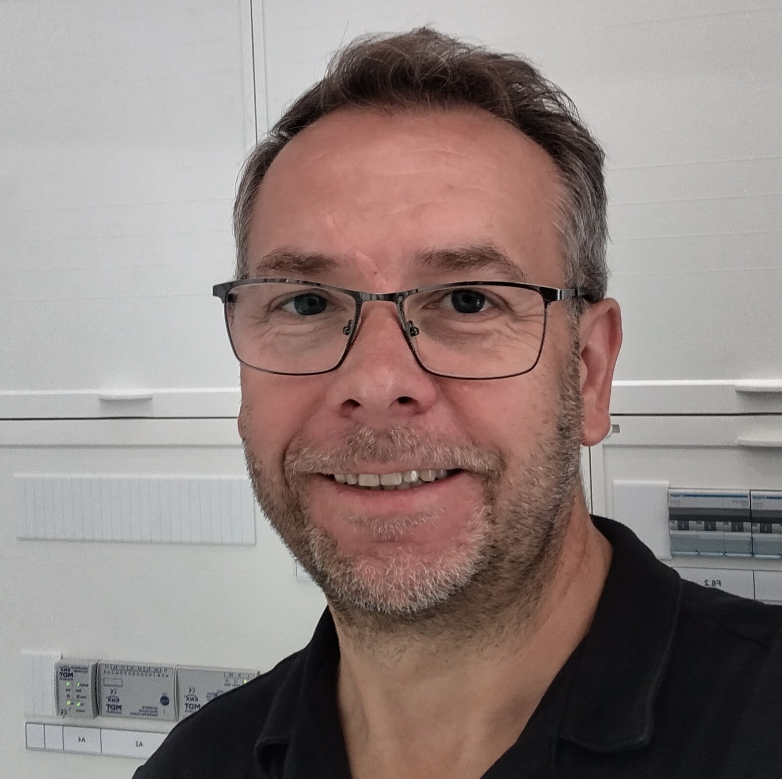 Portret uśmiechniętego elektryka w okularach na tle szafy rozdzielczej z widocznymi bezpiecznikami i modułami sterującymi.