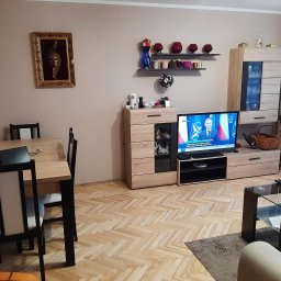 Stanisław Surowczyk - Jasny parkiet ułożony w jodełkę w salonie w Krakowie, widok na stół z krzesłami, szafkę RTV i telewizor z włączonymi wiadomościami.