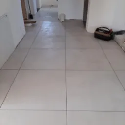 Jasne, kwadratowe płytki podłogowe ułożone w nowym pomieszczeniu, widoczna skrzynka z narzędziami i fragmenty niezakończonych prac wykończeniowych ścian.