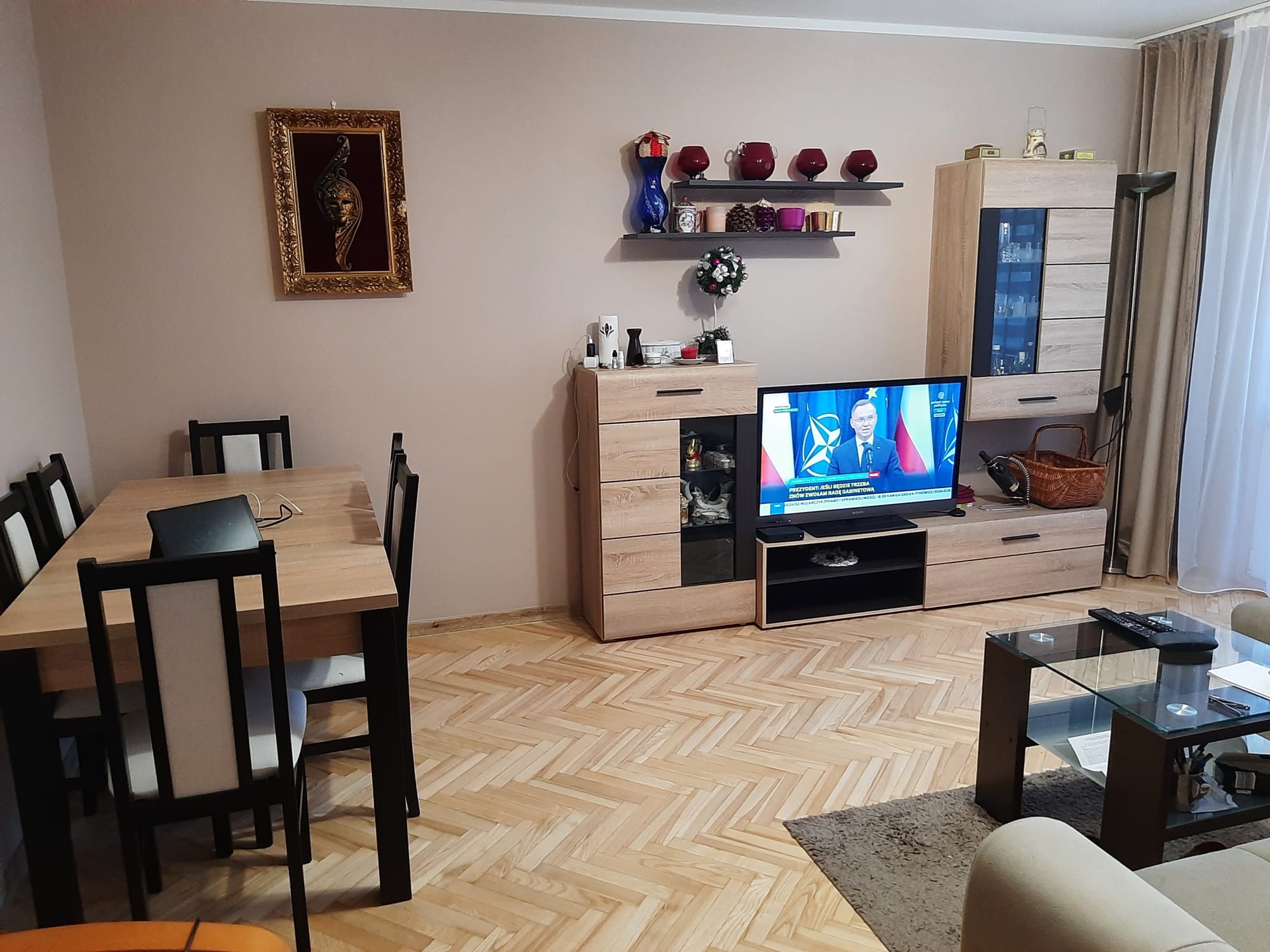 Jasny parkiet ułożony w jodełkę w salonie w Krakowie, widok na stół z krzesłami, szafkę RTV i telewizor z włączonymi wiadomościami.