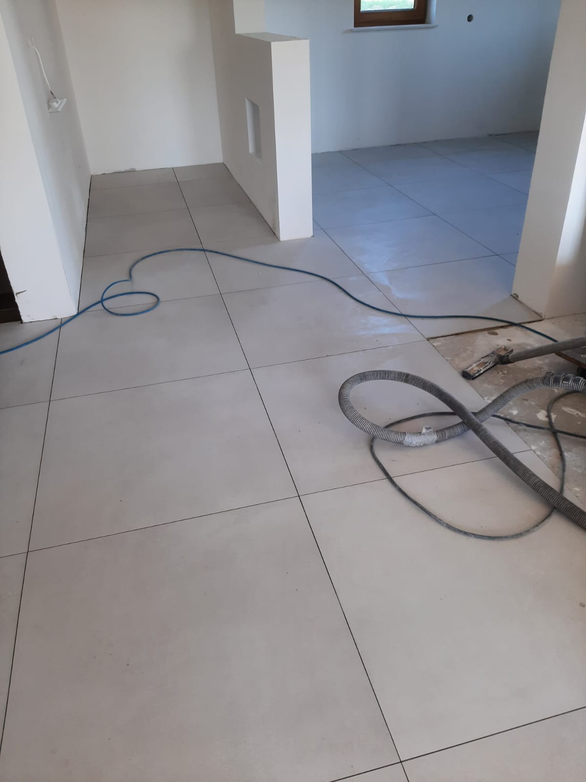 Ułożone, jasne płytki podłogowe z widocznymi fugami w nowym wnętrzu, fragment węża od odkurzacza przemysłowego leżący na podłodze.