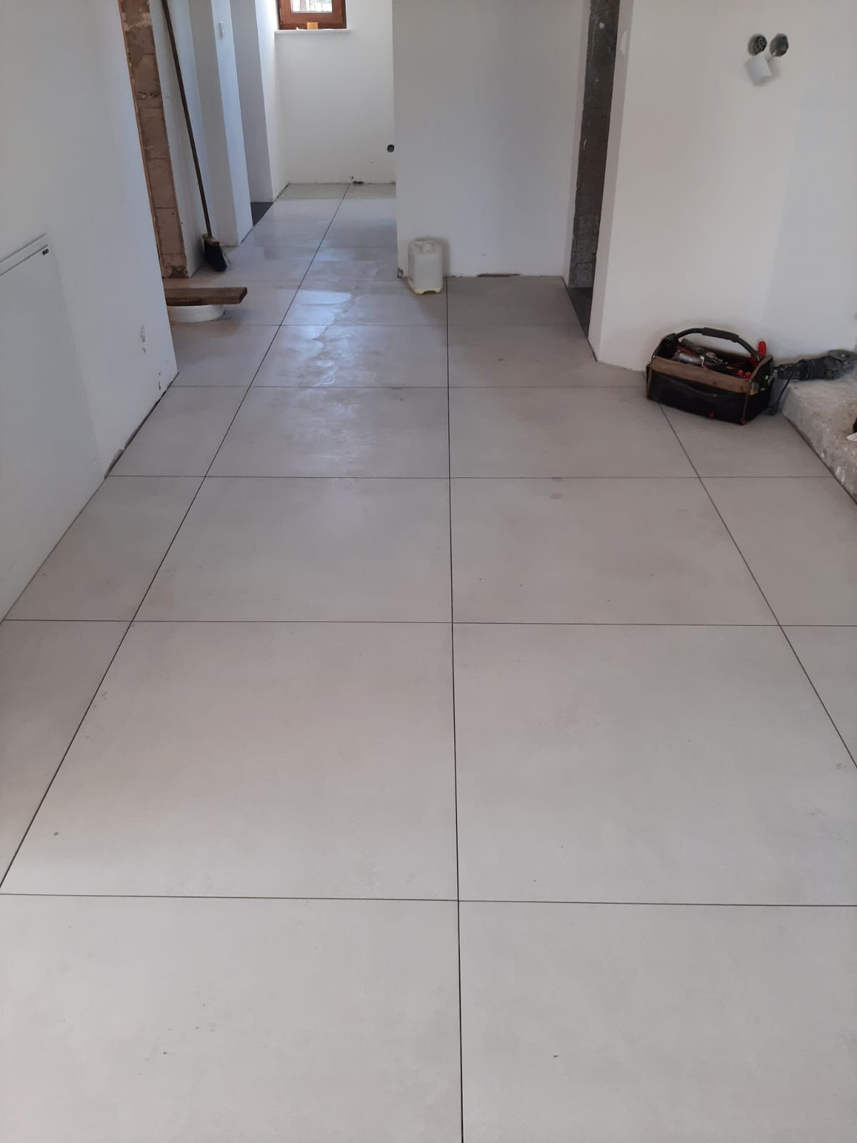 Jasne, kwadratowe płytki podłogowe ułożone w nowym pomieszczeniu, widoczna skrzynka z narzędziami i fragmenty niezakończonych prac wykończeniowych ścian.