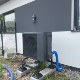 Szara pompa ciepła Panasonic Aquarea zamontowana na betonowych podstawach przy nowoczesnym domu z szaro-białą elewacją. Widoczny niebieski wąż i fragment ogrodzenia.
