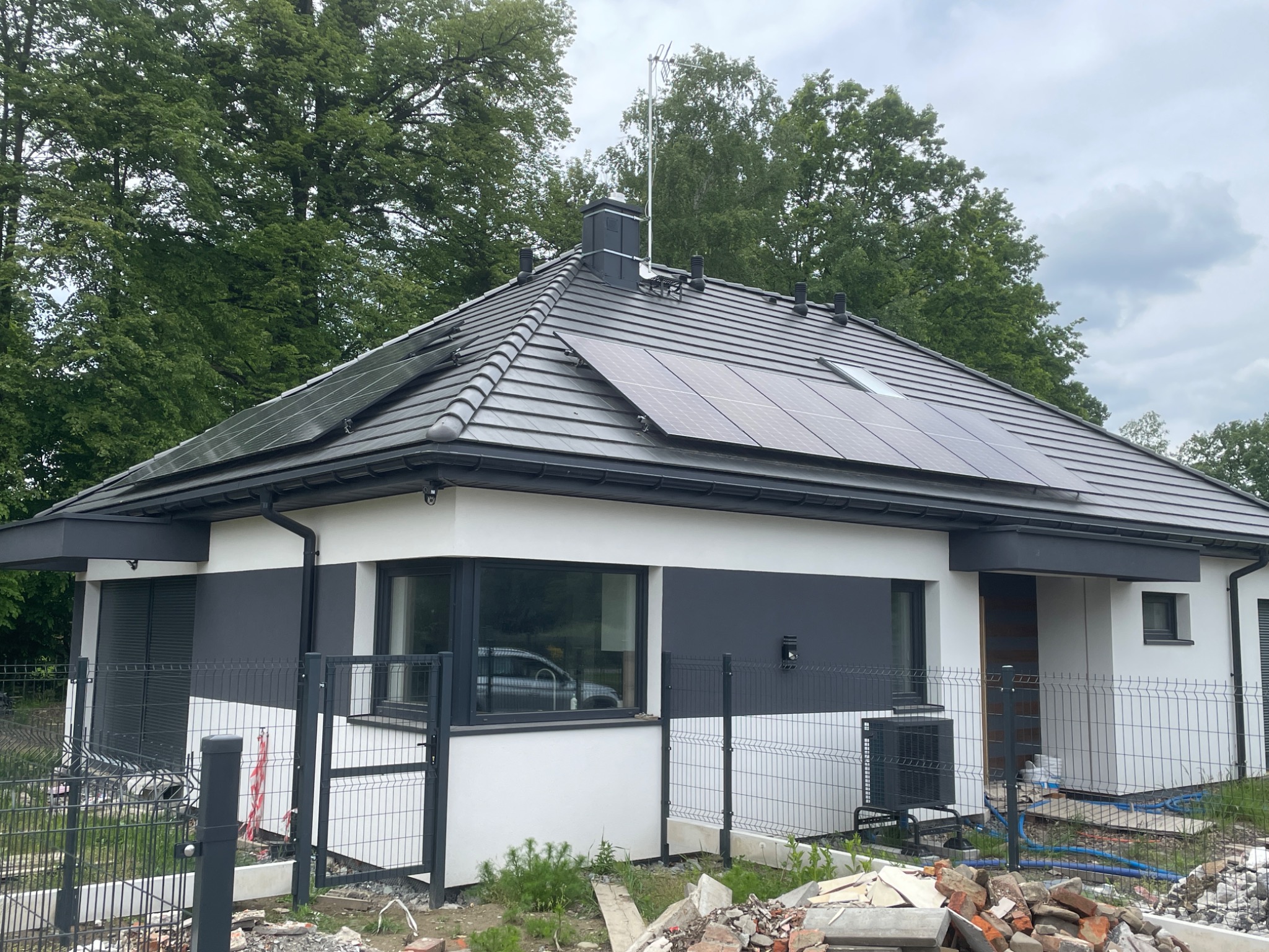 Nowoczesny dom jednorodzinny z ciemną dachówką, panelami słonecznymi i szarą elewacją. Widoczny fragment ogrodzenia panelowego i pompa ciepła przy ścianie budynku.