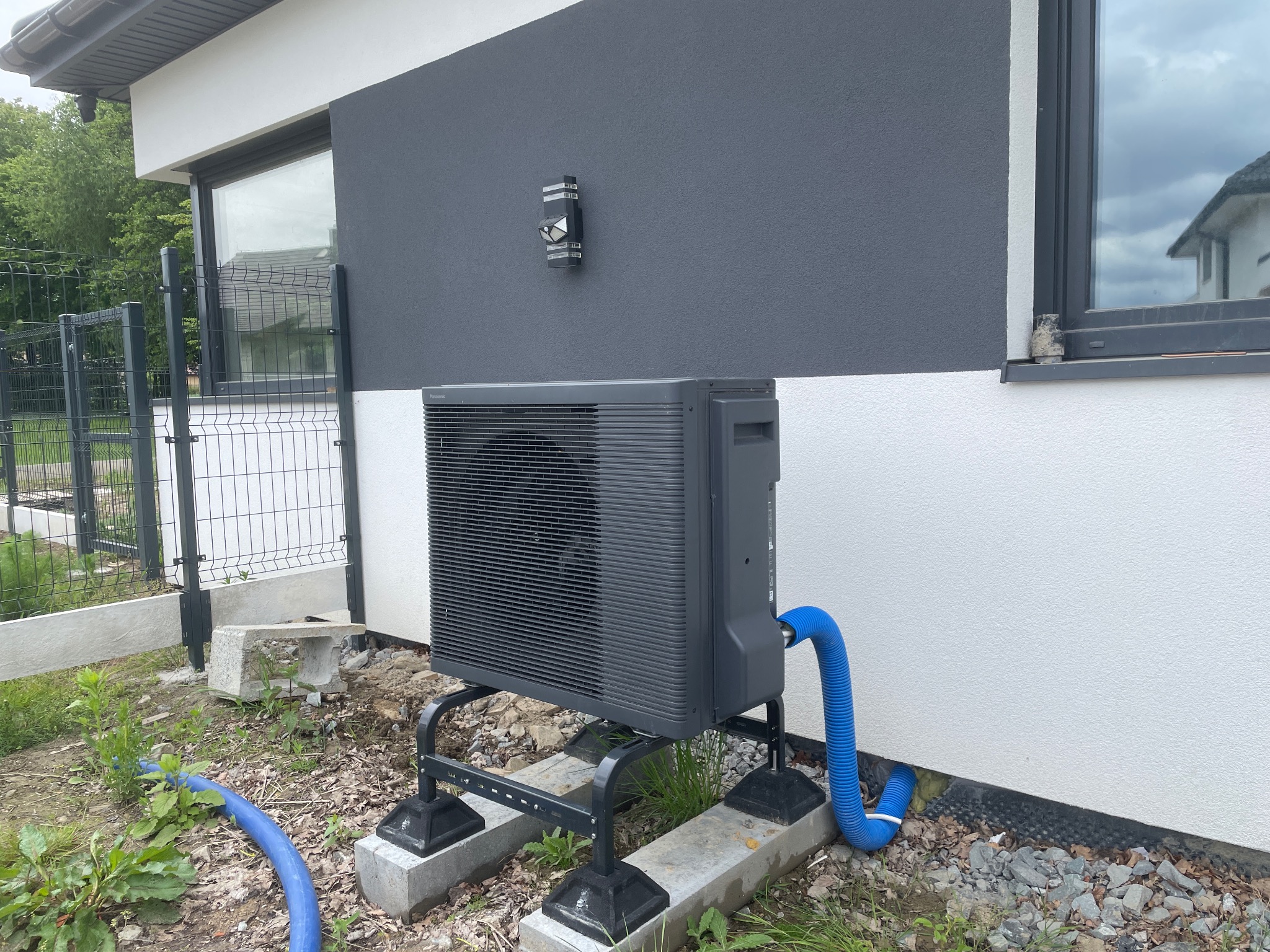 Szara pompa ciepła Panasonic Aquarea zamontowana na betonowych podstawach przy nowoczesnym domu z szaro-białą elewacją. Widoczny niebieski wąż i fragment ogrodzenia.