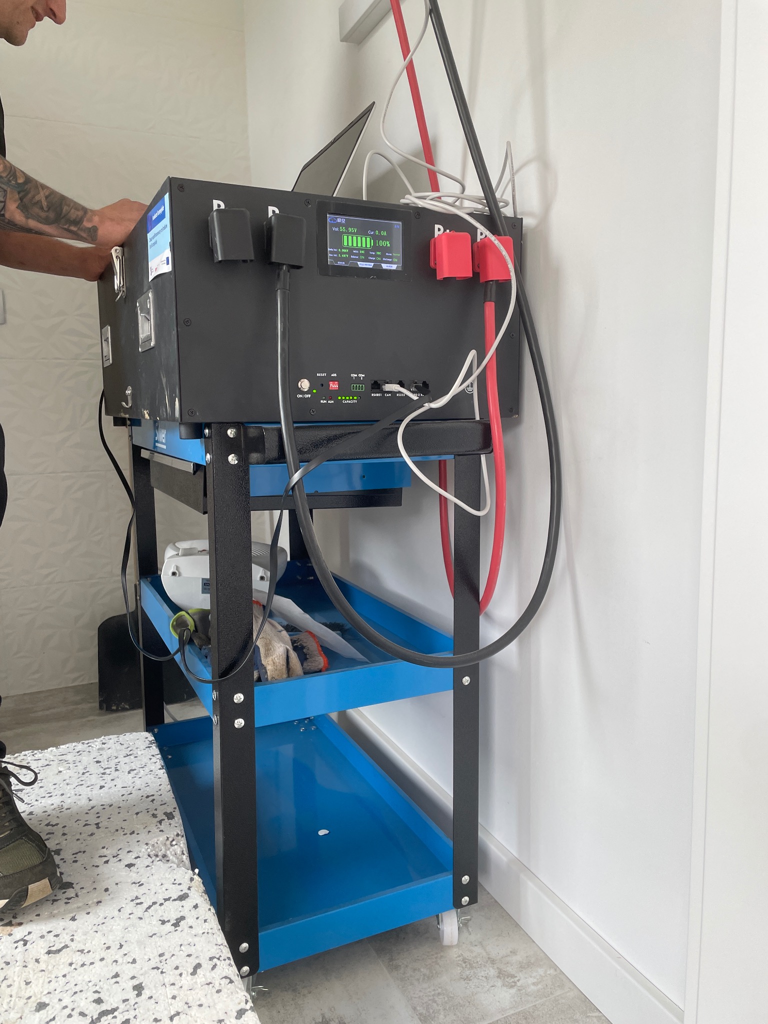 Mobilny magazyn energii na wózku serwisowym, podłączony do instalacji. Widoczny ekran z parametrami pracy i poziomem naładowania baterii. Instalator w trakcie podłączania.