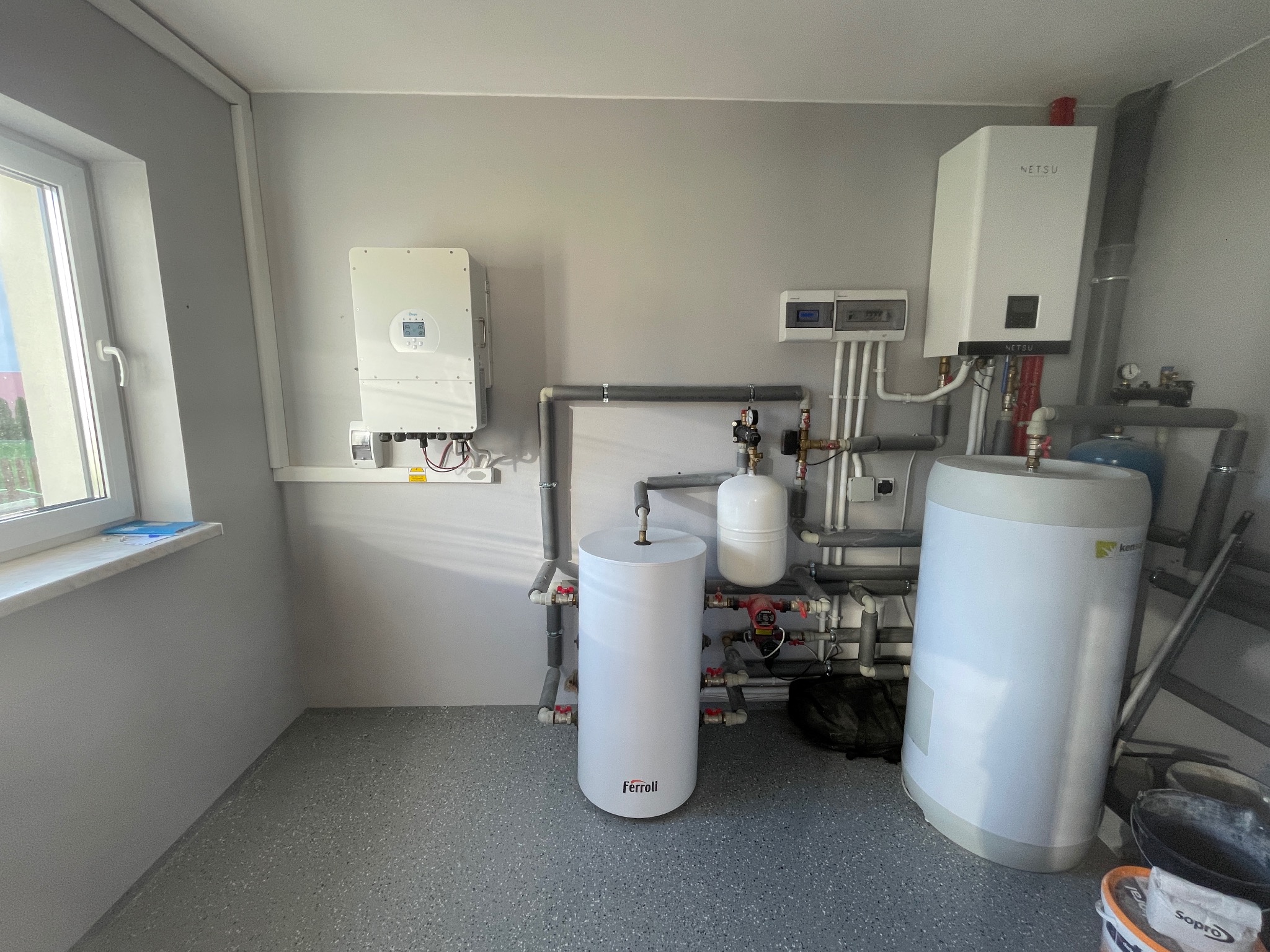 Kompletna instalacja grzewcza w kotłowni: zbiorniki Ferroli i Kensol, pompa ciepła Netsu, inwerter fotowoltaiczny. Szare rury i armatura. Widok na okno.