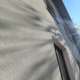 Renova Lublin - Jasna elewacja budynku z widoczną fakturą, oświetlona słońcem, z oknami w ciemnych ramach i widocznymi cieniami drzew.