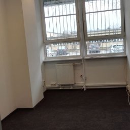 Renova Lublin - Puste pomieszczenie biurowe z ciemną wykładziną, białymi ścianami, grzejnikiem i oknami z kratami, widok na parking.