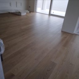 Renova Lublin - Nowo ułożona, jasna podłoga z paneli drewnopodobnych w minimalistycznym, jasnym wnętrzu. W tle widoczne okno balkonowe i przygotowane do dalszych prac wykończeniowych materiały budowlane.