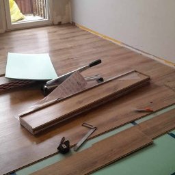 Renova Lublin - Fragment pokoju w trakcie montażu podłogi z paneli drewnopodobnych, widoczne podłoże z zielonej pianki mocowane niebieską taśmą, narzędzia do cięcia i montażu paneli oraz przycięte deski.